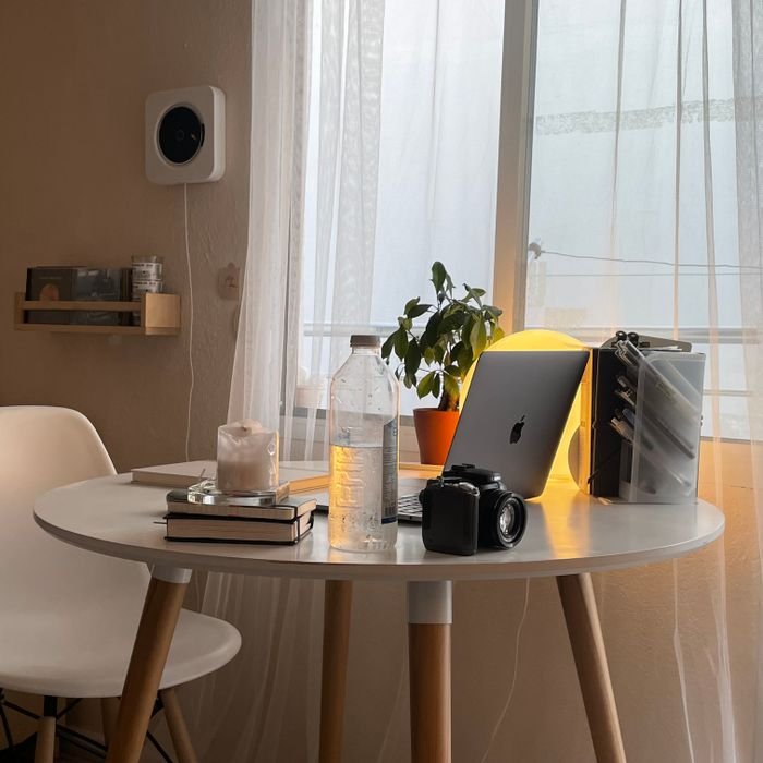 A cozy home workspace featuring a laptop, books, and a camera on a round table by the window.